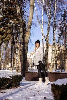 A beautiful young woman in a white jacket and white leather boots walks in a snowy park