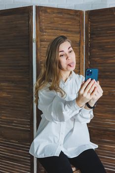 Emotional young woman in a white cotton shirt gesticulates and looks at the screen of a modern gadget. A girl with a mobile phone takes a selfie
