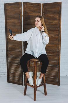Blonde woman with long hair in a white shirt and black pants posing at the phone camera, holding the phone and taking a selfie on an isolated background.