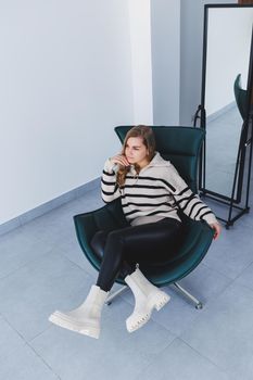 A young beautiful woman is sitting in a chair, wearing white leather boots from a new collection of shoes for spring.
