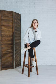 A beautiful woman is wearing a white cotton shirt and black pants. Cheerful emotional girl sits on a wooden chair against a white brick wall.
