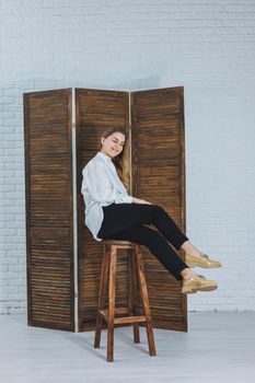 Happy beautiful young woman with a smile in a fashionable outfit with a white shirt, leather beige shoes and black trousers sits on a wooden chair.