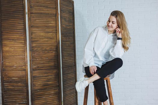 A beautiful woman is wearing a white cotton shirt and black pants. Cheerful emotional girl sits on a wooden chair against a white brick wall.