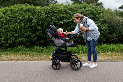 a young mother with a cup of coffee in her hands with a smile on her face straightens the nipple while walking in the park to her little baby in a stroller.