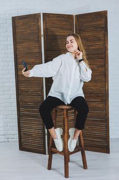 Blonde woman with long hair in a white shirt and black pants posing at the phone camera, holding the phone and taking a selfie on an isolated background.