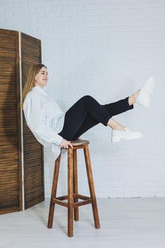 A beautiful woman is wearing a white cotton shirt and black pants. Cheerful emotional girl sits on a wooden chair against a white brick wall.