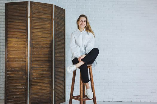 A beautiful woman is wearing a white cotton shirt and black pants. Cheerful emotional girl sits on a wooden chair against a white brick wall.