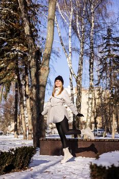A beautiful young woman in a white jacket and white leather boots walks in a snowy park