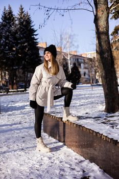 A beautiful young woman in a white jacket and white leather boots walks in a snowy park