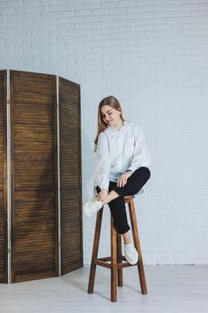 A beautiful woman is wearing a white cotton shirt and black pants. Cheerful emotional girl sits on a wooden chair against a white brick wall.