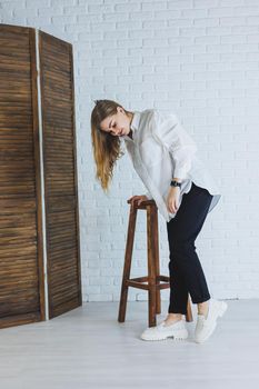 A young woman in black pants and a white shirt against a white brick wall, a stylish modern girl in white leather loafers