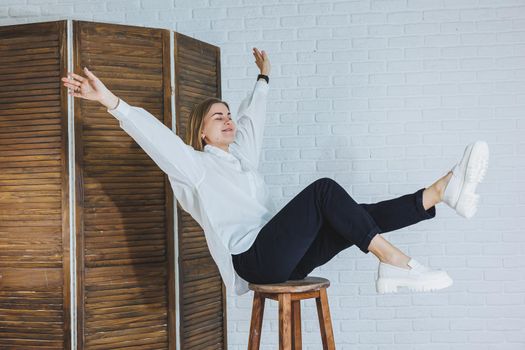 A beautiful woman is wearing a white cotton shirt and black pants. Cheerful emotional girl sits on a wooden chair against a white brick wall.