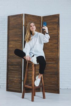 Blonde woman with long hair in a white shirt and black pants posing at the phone camera, holding the phone and taking a selfie on an isolated background.