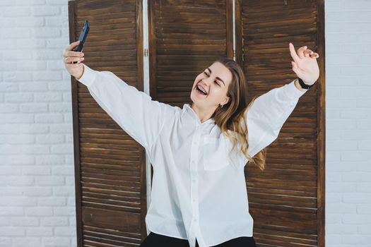 Emotional young woman in a white cotton shirt gesticulates and looks at the screen of a modern gadget. A girl with a mobile phone takes a selfie