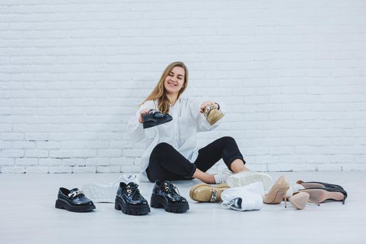 Happy beautiful young woman with a smile in a fashionable outfit with a white shirt and black trousers sits on the floor among the shoes and chooses a new pair of shoes for herself