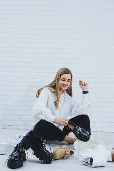 A young woman sits on the floor among shoes and chooses new shoes for herself. Lady in a white shirt and black trousers. Woman in white shoes.