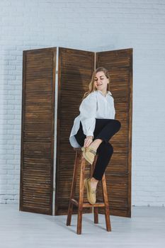 Happy beautiful young woman with a smile in a fashionable outfit with a white shirt, leather beige shoes and black trousers sits on a wooden chair.