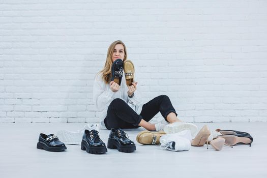 Happy beautiful young woman with a smile in a fashionable outfit with a white shirt and black trousers sits on the floor among the shoes and chooses a new pair of shoes for herself