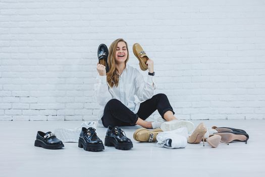 Happy beautiful young woman with a smile in a fashionable outfit with a white shirt and black trousers sits on the floor among the shoes and chooses a new pair of shoes for herself