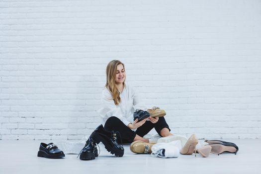Happy beautiful young woman with a smile in a fashionable outfit with a white shirt and black trousers sits on the floor among the shoes and chooses a new pair of shoes for herself