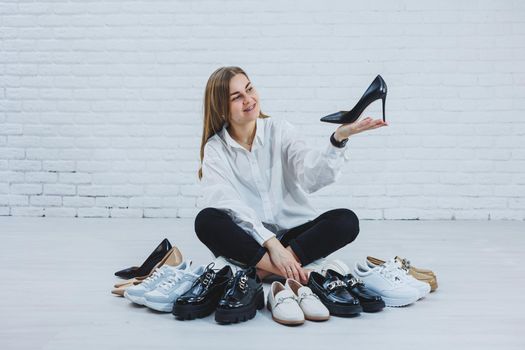 Stylish blonde choosing shoes while sitting on the floor. Side view of a lot of women's shoes on the floor, a choice of stylish shoes.