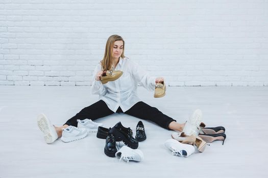 A woman sits on the floor in a dressing room between shoes and chooses new shoes for herself. Lady in a white shirt. Woman in white shoes.