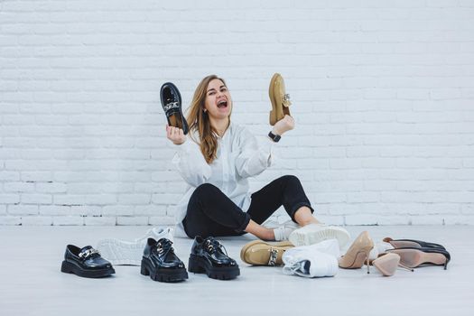 Happy beautiful young woman with a smile in a fashionable outfit with a white shirt and black trousers sits on the floor among the shoes and chooses a new pair of shoes for herself