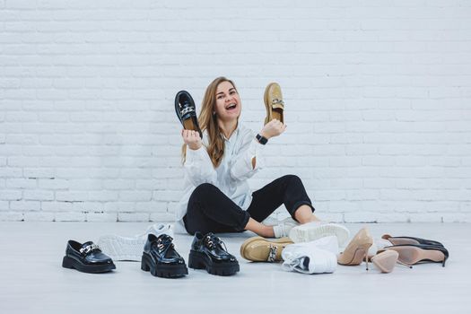 Happy beautiful young woman with a smile in a fashionable outfit with a white shirt and black trousers sits on the floor among the shoes and chooses a new pair of shoes for herself