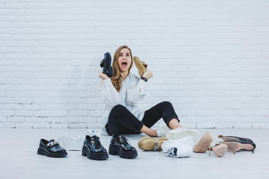 Happy beautiful young woman with a smile in a fashionable outfit with a white shirt and black trousers sits on the floor among the shoes and chooses a new pair of shoes for herself