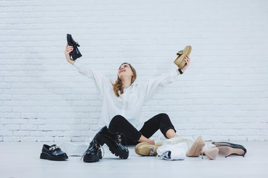 Happy beautiful young woman with a smile in a fashionable outfit with a white shirt and black trousers sits on the floor among the shoes and chooses a new pair of shoes for herself