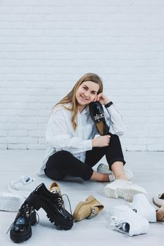 A young woman sits on the floor among shoes and chooses new shoes for herself. Lady in a white shirt and black trousers. Woman in white shoes.