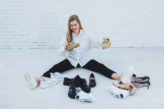 A woman sits on the floor in a dressing room between shoes and chooses new shoes for herself. Lady in a white shirt. Woman in white shoes.