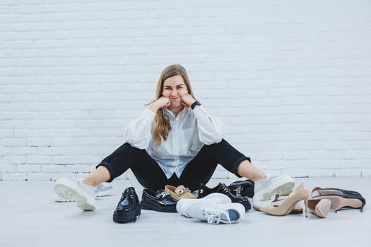 Happy beautiful young woman with a smile in a fashionable outfit with a white shirt and black trousers sits on the floor among the shoes and chooses a new pair of shoes for herself