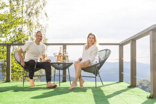 Happy couple sit on wicker chairs at outdoor terrace in the mountains and drink wine. Romantic time together