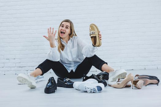 Stylish blonde choosing shoes while sitting on the floor. Side view of a lot of women's shoes on the floor, a choice of stylish shoes.