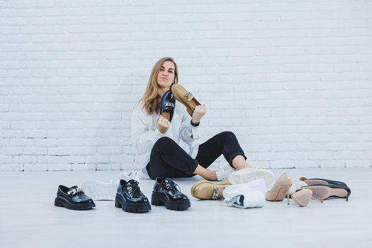 Happy beautiful young woman with a smile in a fashionable outfit with a white shirt and black trousers sits on the floor among the shoes and chooses a new pair of shoes for herself