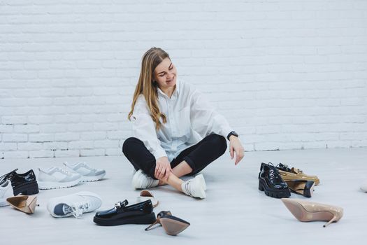 A young woman sits on the floor among shoes and chooses new shoes for herself. Lady in a white shirt and black trousers. Woman in white shoes.