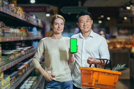 Young family diverse couple of shoppers in supermarket, smiling and looking at camera, grocery department, man and woman holding shopping basket and showing green screen of smartphone.