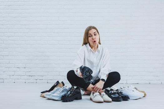 Happy beautiful young woman with a smile in a fashionable outfit with a white shirt and black trousers sits on the floor among the shoes and chooses a new pair of shoes for herself