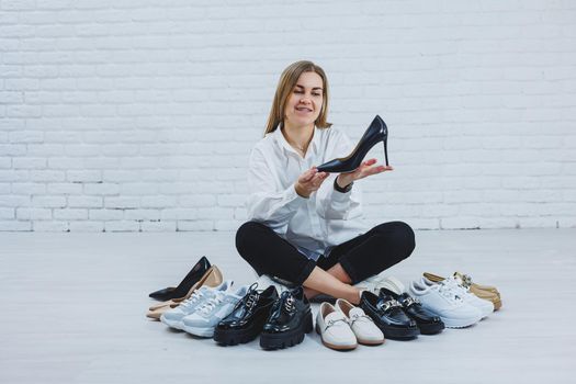 Stylish blonde choosing shoes while sitting on the floor. Side view of a lot of women's shoes on the floor, a choice of stylish shoes.
