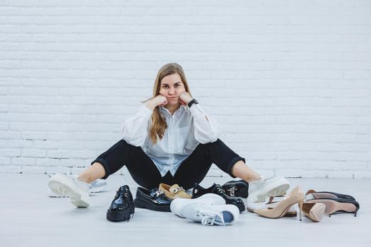 Stylish blonde choosing shoes while sitting on the floor. Side view of a lot of women's shoes on the floor, a choice of stylish shoes.