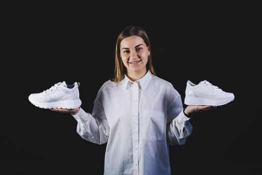 A fair-haired woman in a white shirt on a black background holds white sneakers made of genuine leather in her hands