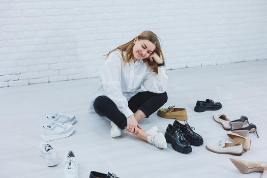 A young woman sits on the floor among shoes and chooses new shoes for herself. Lady in a white shirt and black trousers. Woman in white shoes.