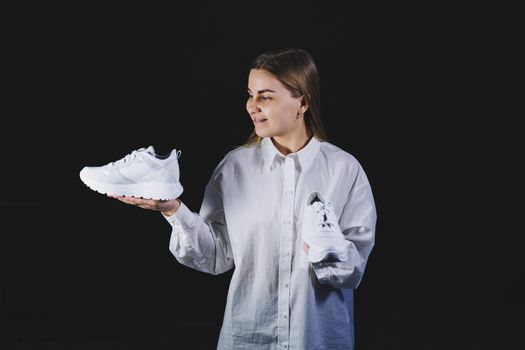 A fair-haired woman in a white shirt on a black background holds white sneakers made of genuine leather in her hands