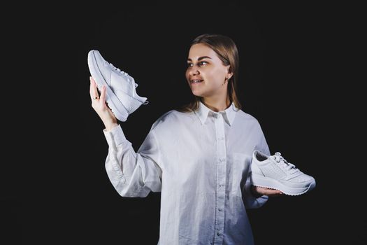 A fair-haired woman in a white shirt on a black background holds white sneakers made of genuine leather in her hands