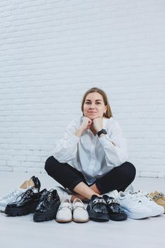 Happy beautiful young woman with a smile in a fashionable outfit with a white shirt and black trousers sits on the floor among the shoes and chooses a new pair of shoes for herself
