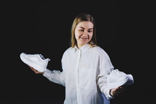 A fair-haired woman in a white shirt on a black background holds white sneakers made of genuine leather in her hands