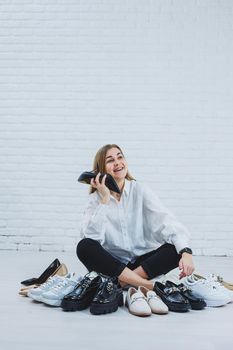 Happy beautiful young woman with a smile in a fashionable outfit with a white shirt and black trousers sits on the floor among the shoes and chooses a new pair of shoes for herself