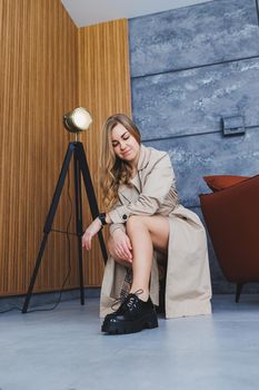 Young beautiful woman sits on the floor in a beige spring raincoat and modern stylish black shoes. Stylish woman of European appearance with long blond hair