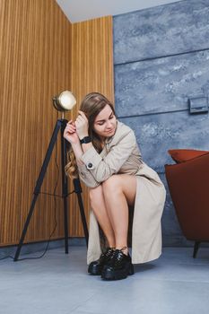 Young beautiful woman sits on the floor in a beige spring raincoat and modern stylish black shoes. Stylish woman of European appearance with long blond hair